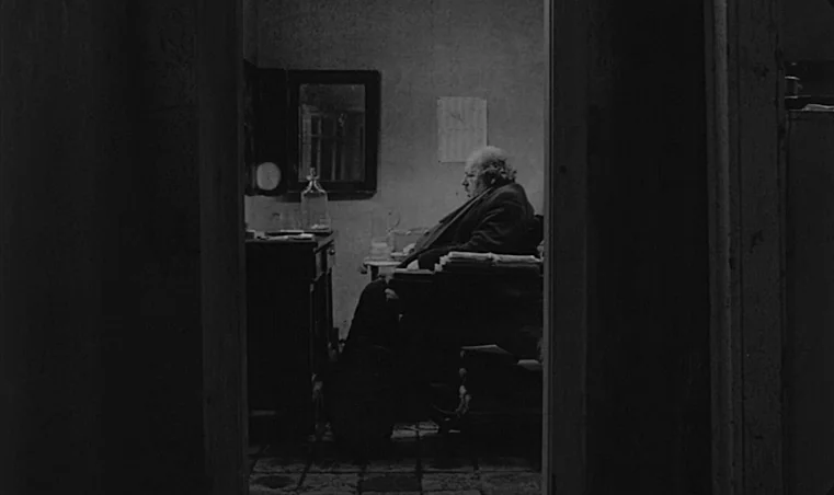 Sátántangó (1994) movie still: medium — A sparsely furnished office contains a heavy desk and a square mirror on the wall. A stout man sits alone at…