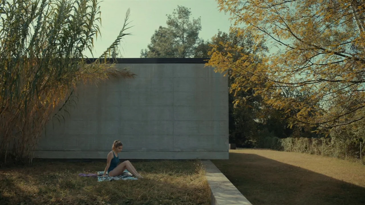 Amanda (2022) movie still: wide — A modernist concrete structure stands in a grassy area surrounded by trees and vegetation. A young woman in a…
