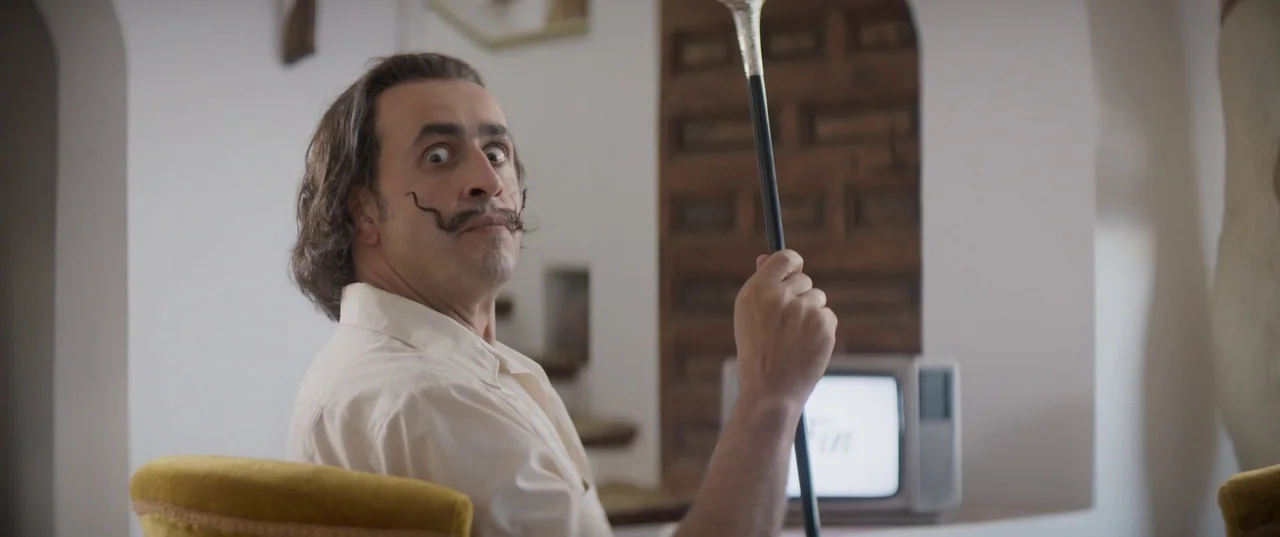 Daaaaaalí! (2023) movie still: medium — A man resembling Salvador Dali sits in a mustard yellow chair. He holds a black walking stick while glancing…