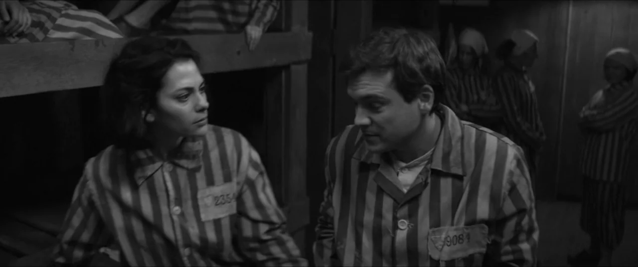Bau, Artist at War (2025) movie still: medium — A dark wooden interior features bunk beds and people wearing thin striped prison uniforms. Two…
