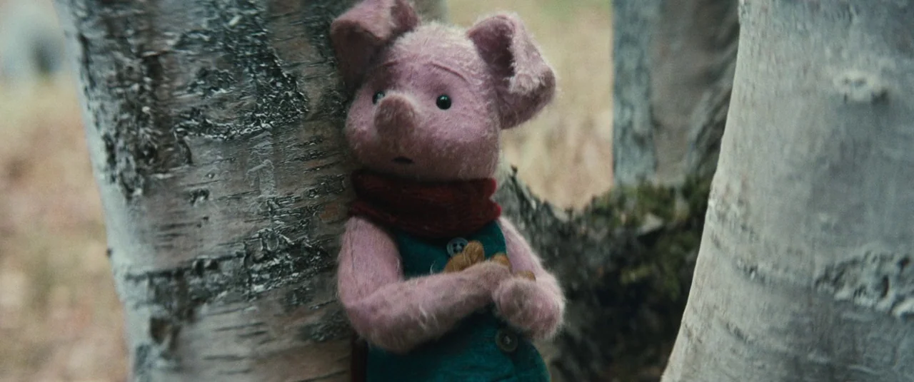 Christopher Robin (2018) movie still: medium — A small pink plush toy stands between two thick birch trees in a woodland. The character holds its…