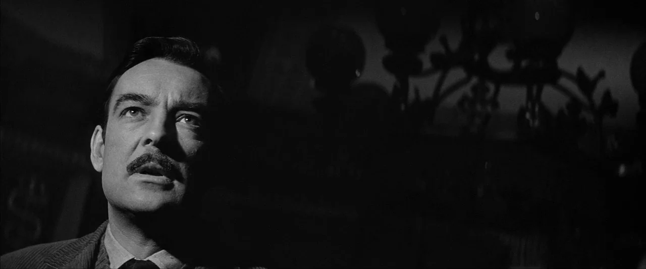 The Haunting (1963) movie still: closeup — A dark, dimly lit space contains a prominent ornate iron chandelier in the background. A man stands in…