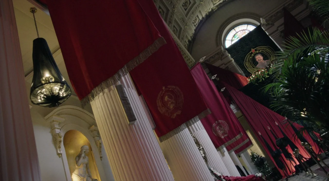 The Death of Stalin (2017) movie still: establishing — A grand neoclassical hall with white fluted pillars displays draped red Soviet banners. A…