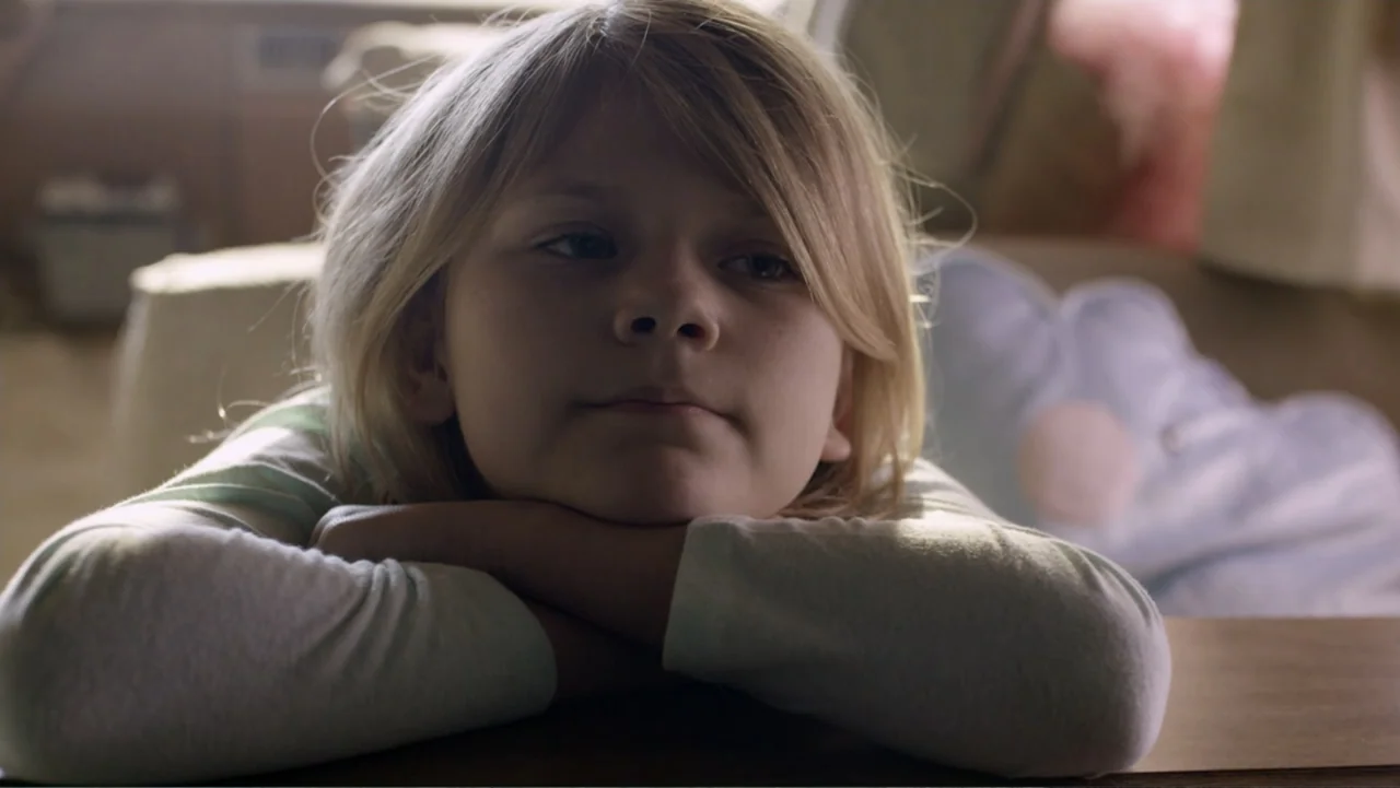 Totem (2022) movie still: closeup — A sunlit living room with a neutral-colored interior space and blurred background elements. A young girl with…