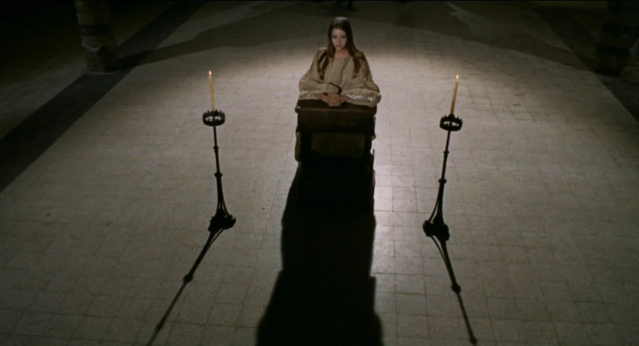 Beatrice Cenci (1969) movie still: wide — A dark stone floor in a cathedral features a woman kneeling at a wooden kneeler. Two iron candle holders…