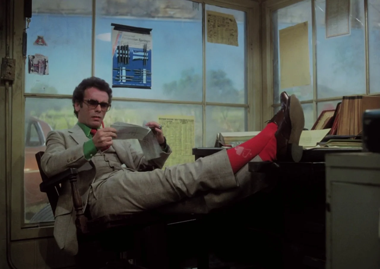 Human Highway (1982) movie still: medium — A cluttered small office has glass walls looking out onto a bright exterior field. A man in a suit sits…