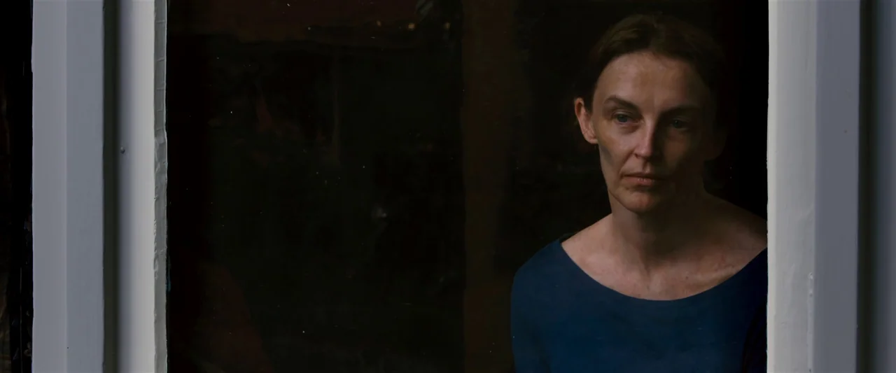 The Loved Ones (2009) movie still: medium — A woman stands in a dark interior looking through a glass window pane. The frame is divided by white…