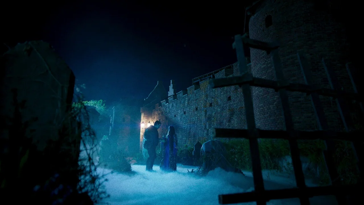 The Munsters (2022) movie still: wide — A stone fortification with high walls stands at night under moonlight. A man and woman converse within a…