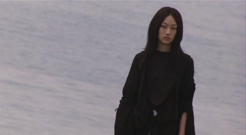 Rampo Noir (2005) movie still: medium — A young woman stands against an expansive, grey-toned water background under an overcast sky. She is…