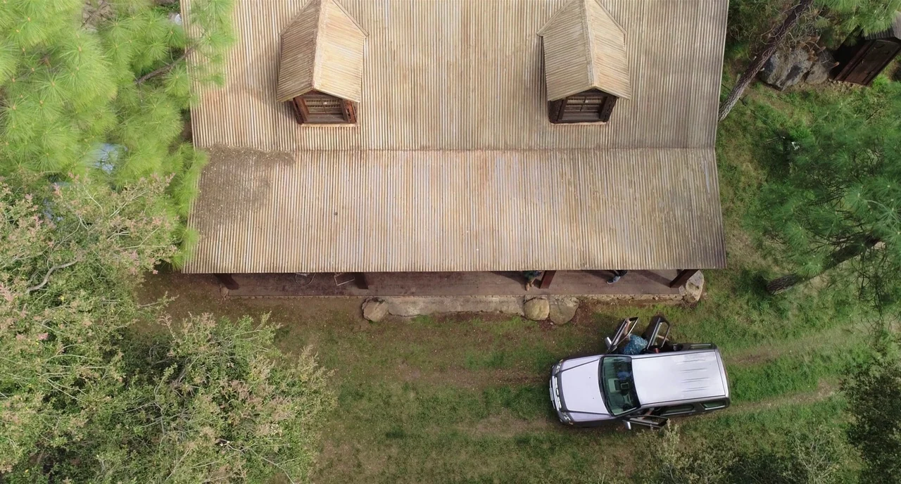 Nightmare Cinema movie still: establishing — A rural cabin features a corrugated metal roof and two small attic dormers. A silver SUV is parked on a…