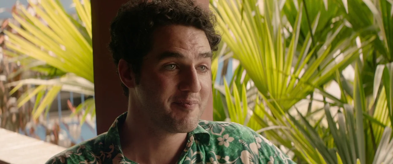 Stars At Noon (2022) movie still: medium — An outdoor patio area is surrounded by lush green palm fronds. A young man with curly hair wears a floral…