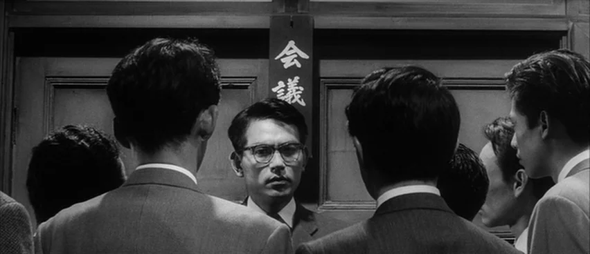 The Bad Sleep Well (1960) movie still: medium — A group of men in suits stand in a dark, wood-paneled room. The men face a central man who is…
