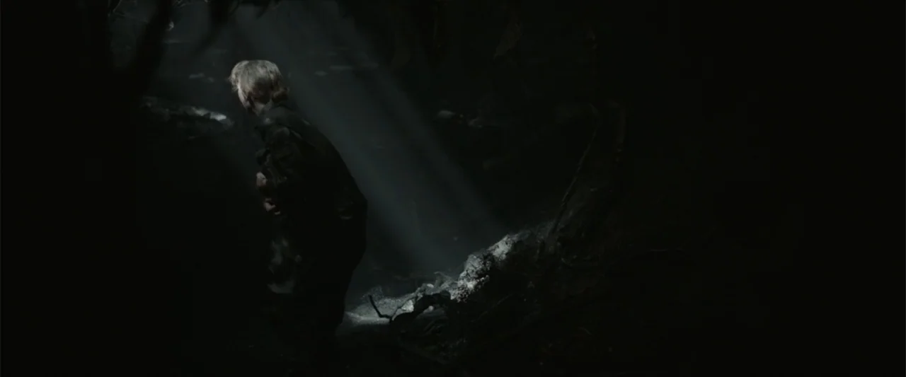 Monster Hunter (2020) movie still: medium — A dark, rocky cave interior is pierced by a sharp, concentrated shaft of sunlight. A man stands…