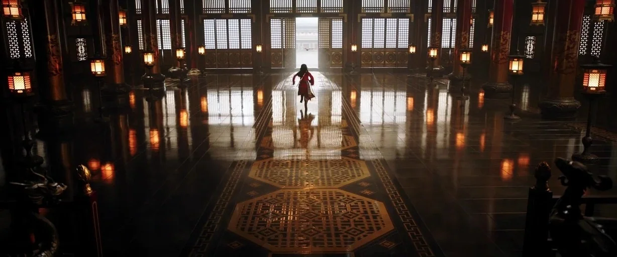 Mulan (2020) movie still: wide — A vast, dark wood-paneled hall features highly reflective floors and rows of ornate lanterns. A small girl in a…