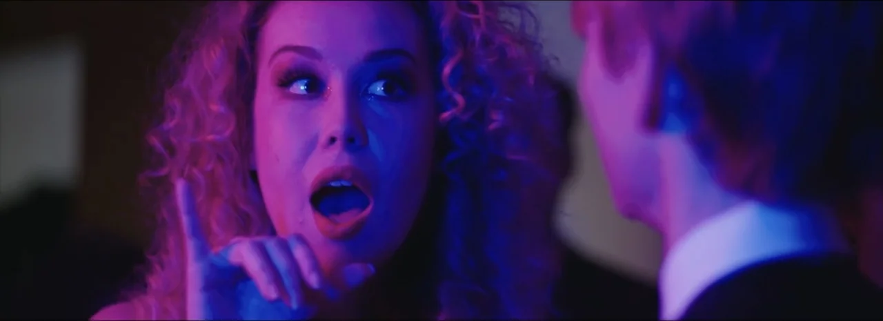 DannyBoy (2020) movie still: closeup — A dark indoor space is filled with intense magenta and blue neon light. A blonde woman speaks to a man in a…