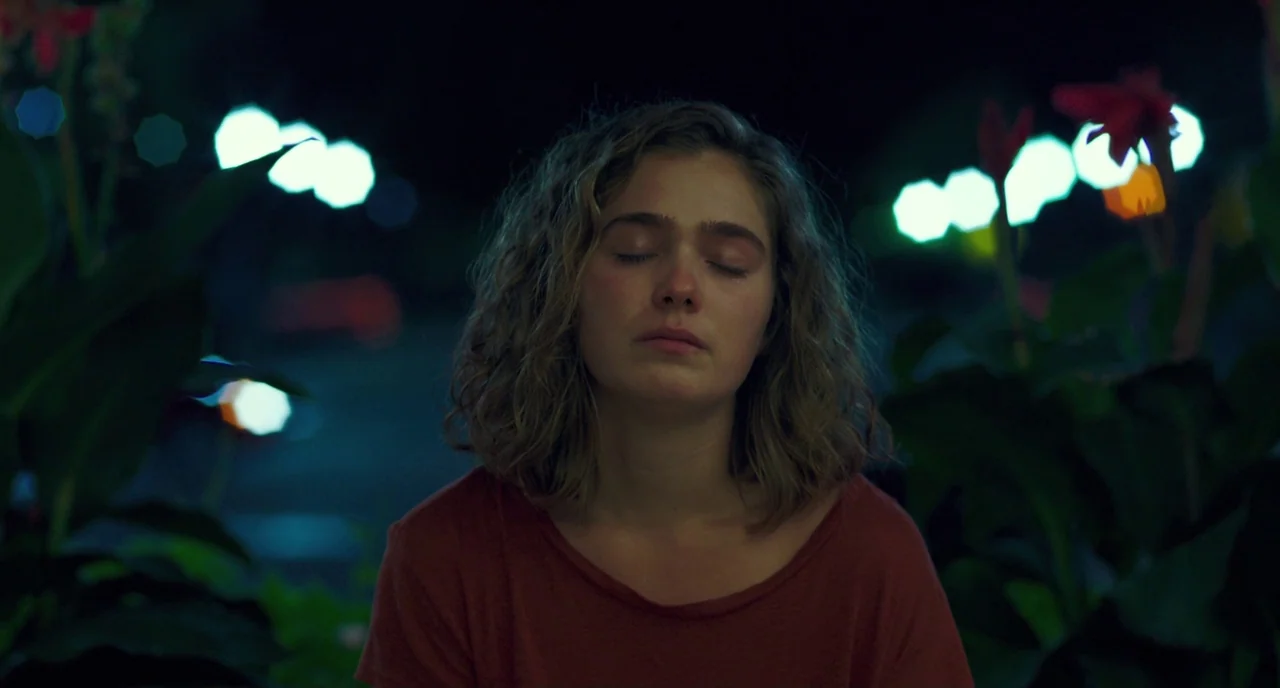 Columbus (2017) movie still: medium — A woman stands outdoors at night framed by dark out-of-focus foliage. She keeps her eyes closed while standing…