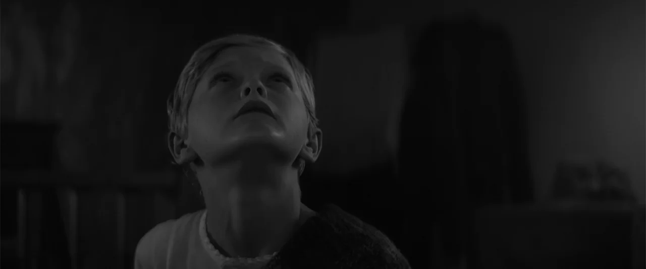 A Christmas Mystery (2022) movie still: closeup — A dark indoor setting features a child in the foreground looking toward an unseen height. The boy…