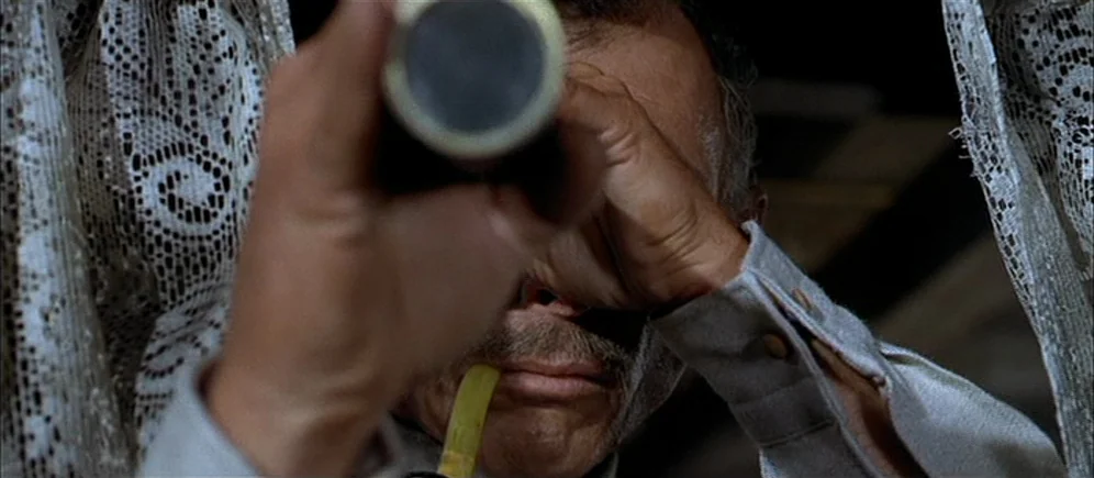 For A Few Dollars More (1965) movie still: closeup — The man stands between white lace curtains while peering through a handheld telescope. He holds…