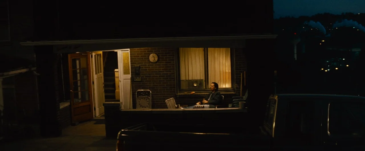Out of the Furnace (2013) movie still: wide — A residential porch at night with brick siding and a bright interior window. A man sits alone in a…