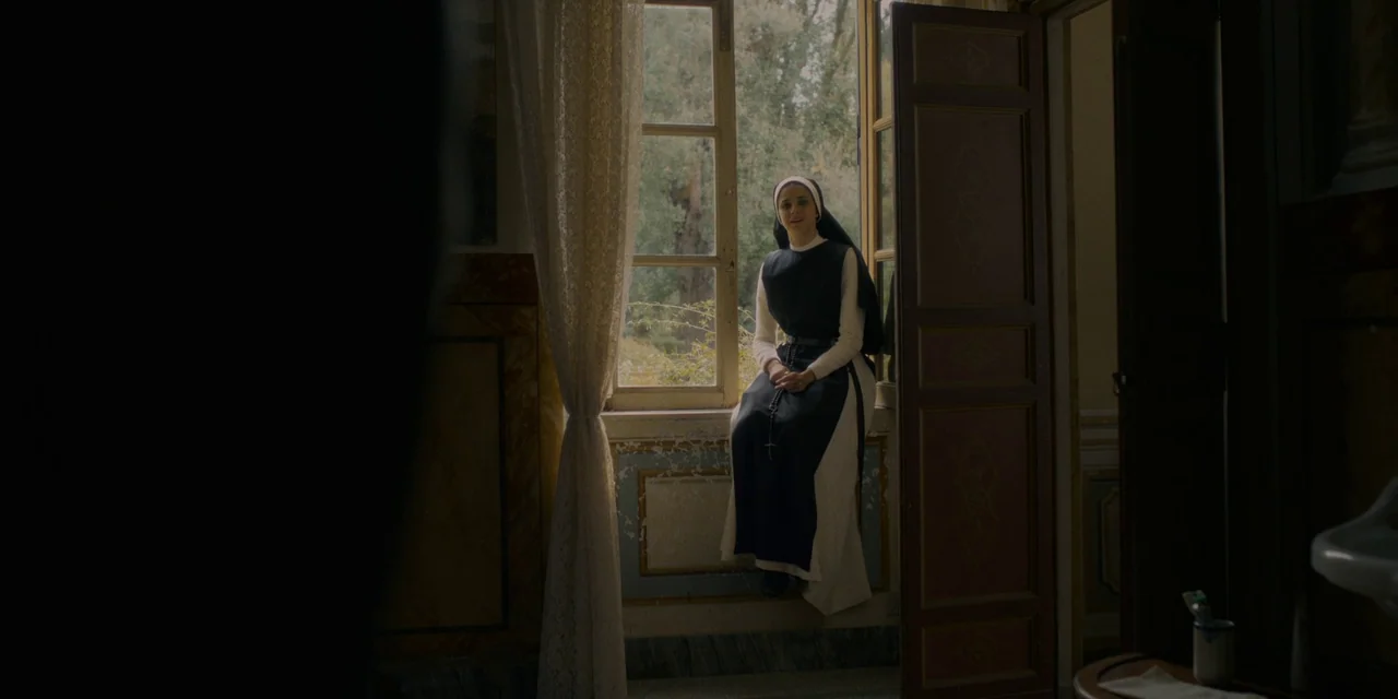 Immaculate (2024) movie still: fullbody — A dimly lit room with an open window looks out onto greenery. A nun in traditional attire sits on the…