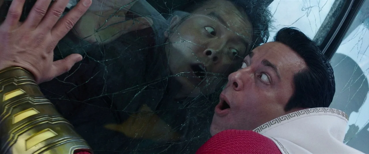 Shazam! (2019) movie still: closeup — A man in a red suit stands behind a person reflected in broken glass. Both individuals press their faces…