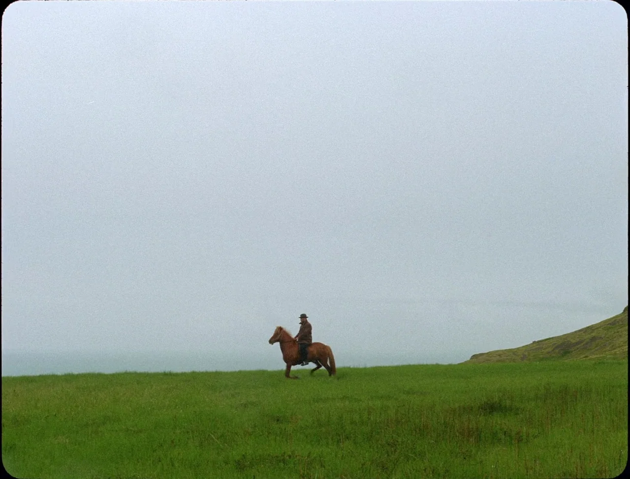 Godland (2022) movie still: wide — A vast, open green meadow stretches under a flat, grey, overcast sky. A man on a brown horse travels slowly…