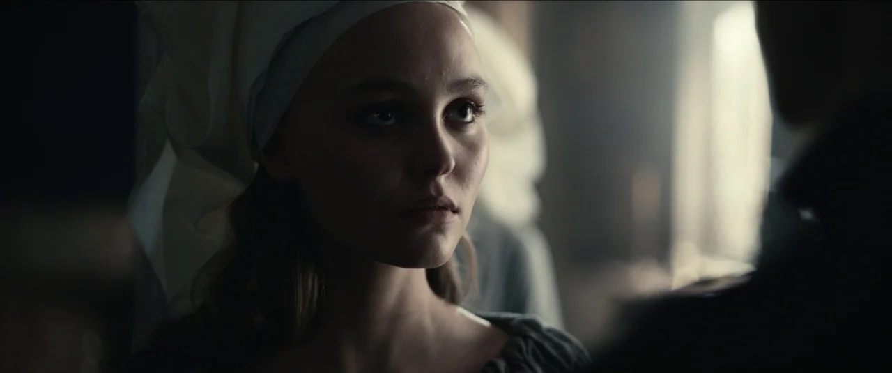 The King movie still: closeup — A dimly lit stone chamber with soft natural light source from a window. A woman wearing a white medieval head…
