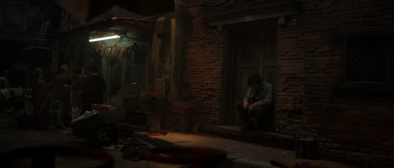 Doctor Strange (2016) movie still: wide — A narrow alleyway with red brick buildings and a small, dimly lit shop stall. A man sits motionless in the…