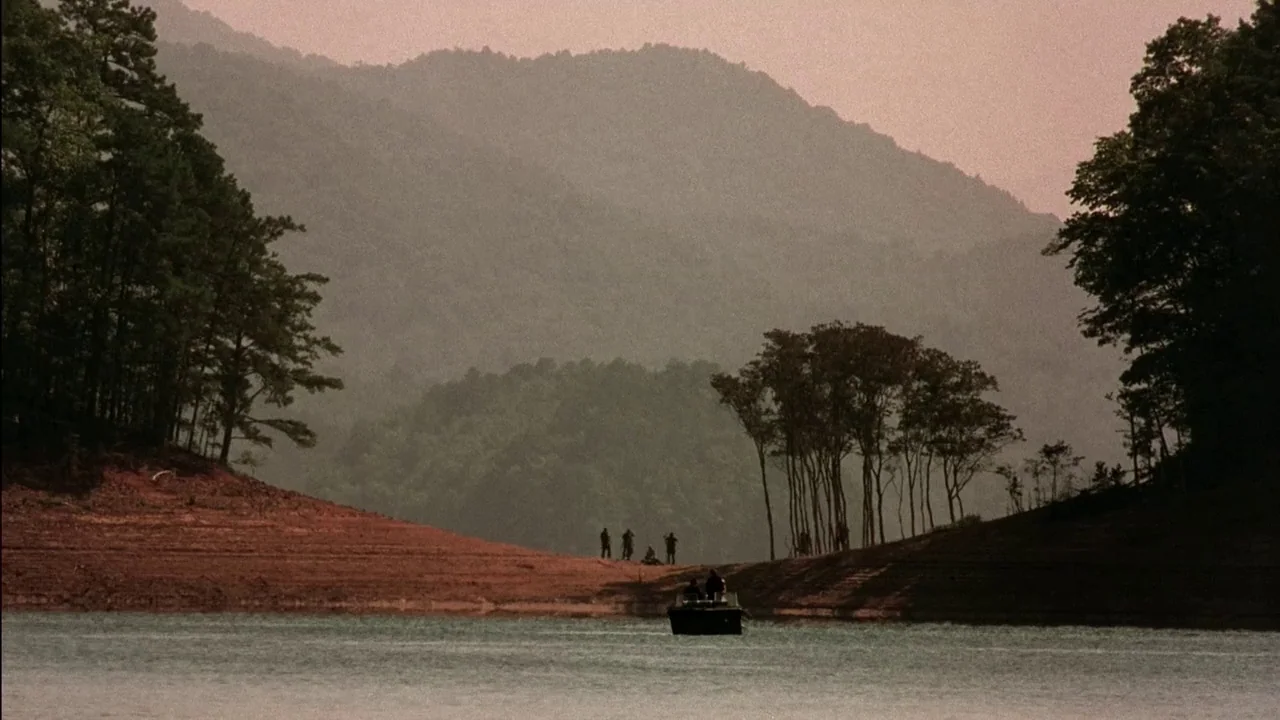 In Dreams (1999) movie still: wide — A wide lake sits between two tree-lined hills under a soft pink sunset sky. A small boat carrying several…