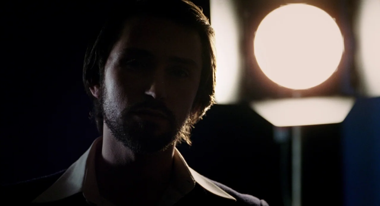 Pasolini (2014) movie still: closeup — A dark, minimally furnished space featuring a single bright round lighting fixture. A man with a beard stands…