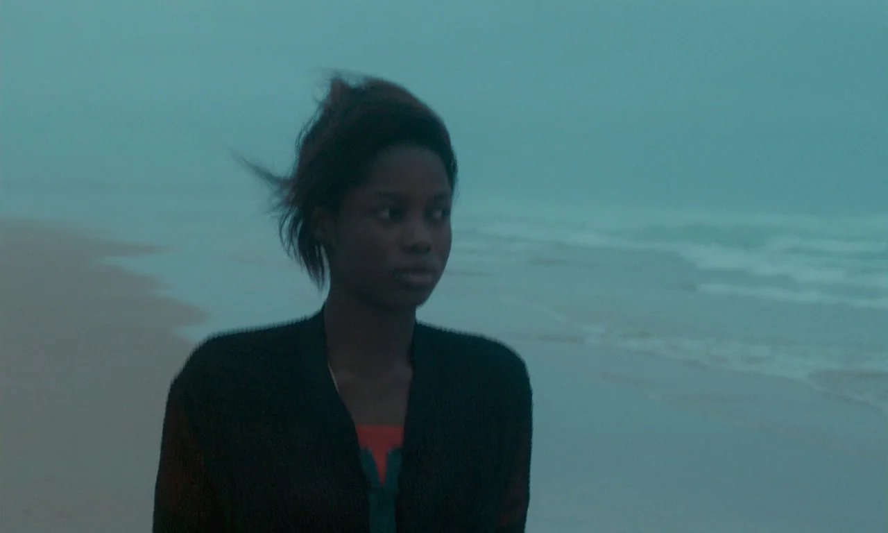 Atlantics (2019) movie still: medium — A woman walks along a misty shoreline under a heavily overcast sky. Waves roll gently onto the sand behind…