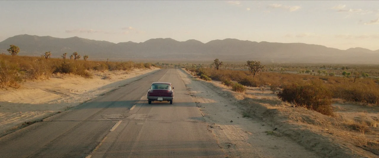 The Last Stop in Yuma County (2023) movie still: wide — A desolate paved road stretches across a dry, arid desert landscape at sunset. A lone…