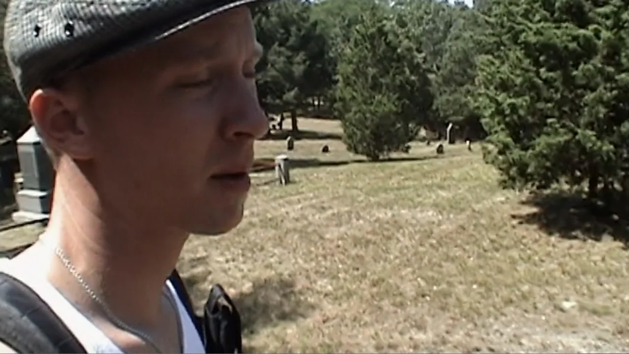 Daniel (1983) movie still: closeup — An open grassy area with scattered headstones and mature trees. A man in a flat cap stands in the foreground…