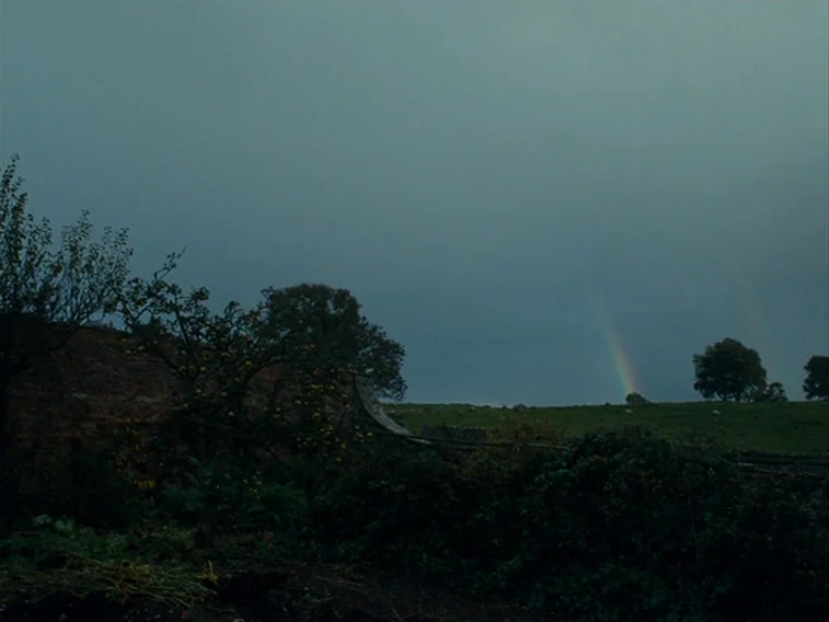Wuthering Heights movie still: establishing — A rural landscape features an old brick wall bordering a green grassy hillside field. A faint rainbow…