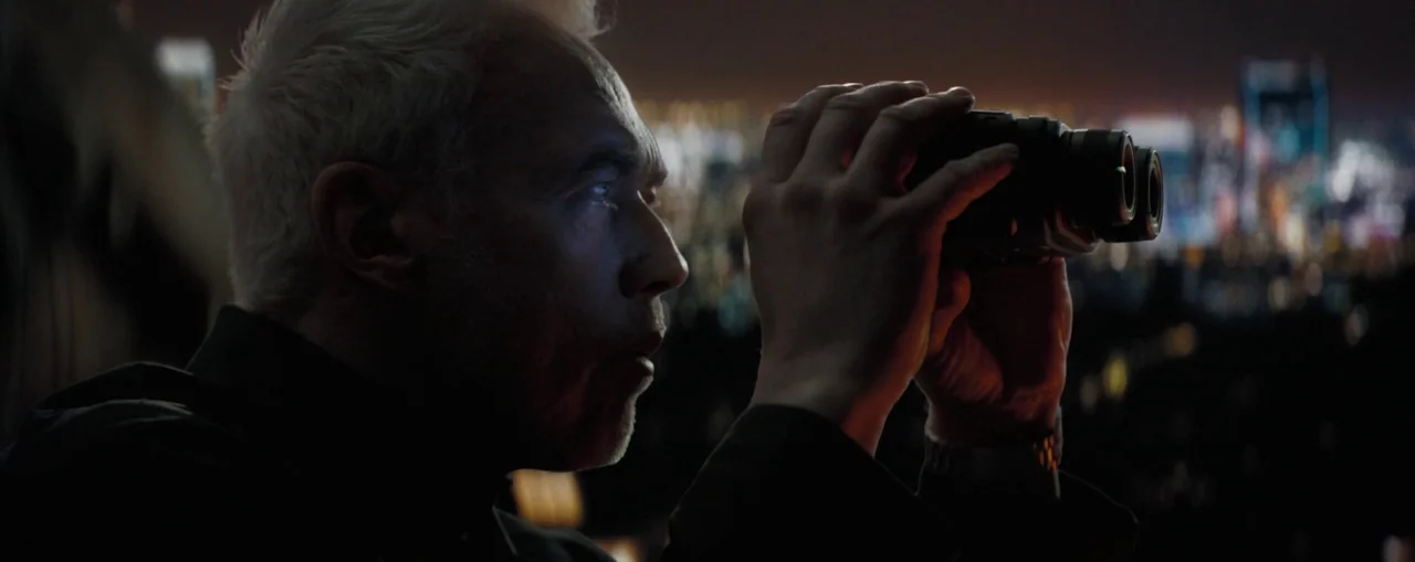 The Naked Gun (2025) movie still: closeup — An elevated urban vantage point looks over a blurry cityscape at night. An elderly man stares through a…