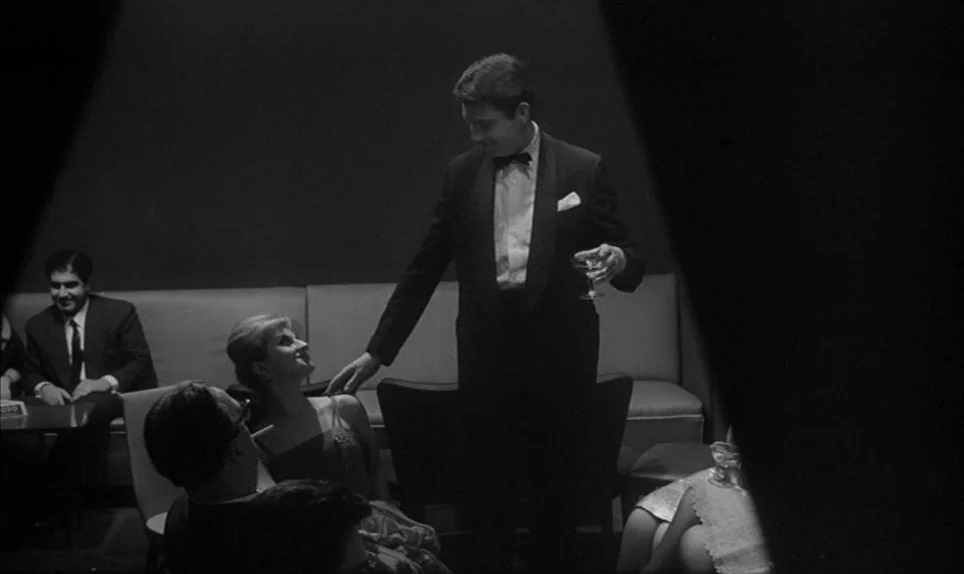 Eva (1962) movie still: medium — A dimly lit lounge with modern minimalist chairs and a long padded bench seat. A man in a tuxedo stands holding a…