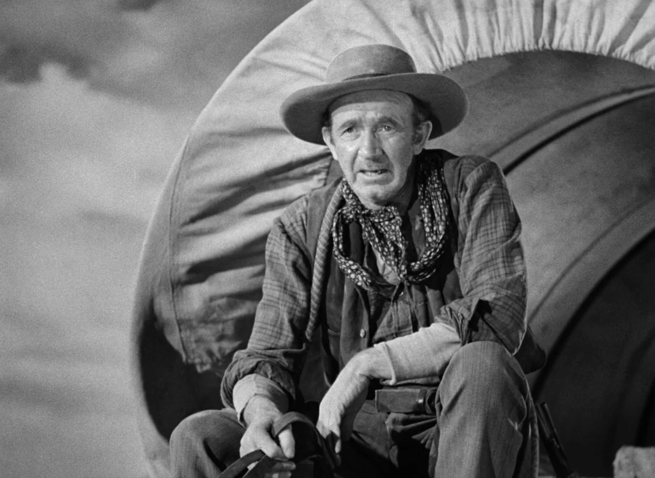 Red River (1948) movie still: medium — A man sits on the back of a canvas-covered wagon in an open landscape. The subject holds leather reins while…