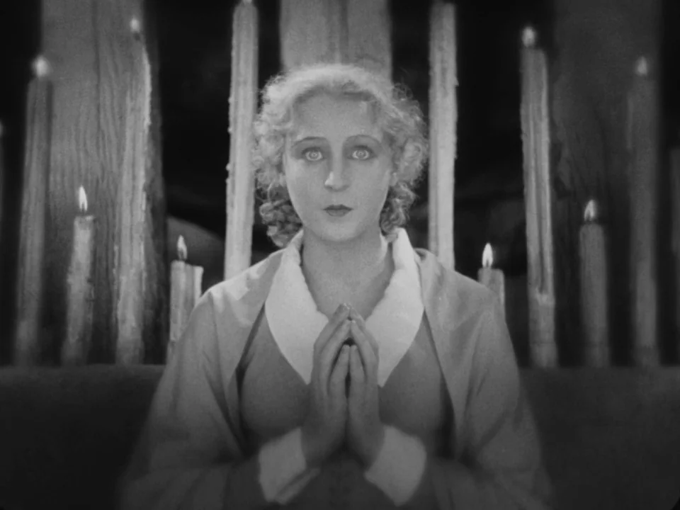Metropolis (1927) movie still: closeup — A woman stands before a row of upright candles in a dim space. She presses her palms together in a prayer…