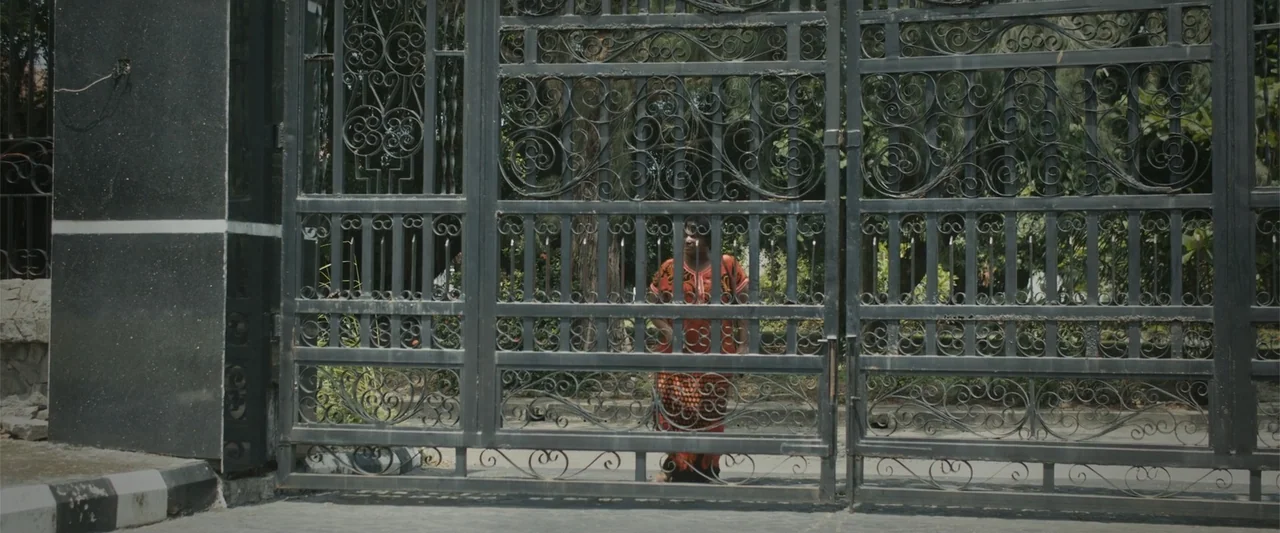 Casa De Novia (2024) movie still: wide — A tall, black wrought-iron gate stands before a residential property with lush trees behind. A woman in a…