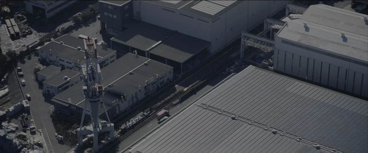 Kate (2021) movie still: establishing — An aerial view displays a complex of large metal industrial warehouses and buildings. A tall white crane…