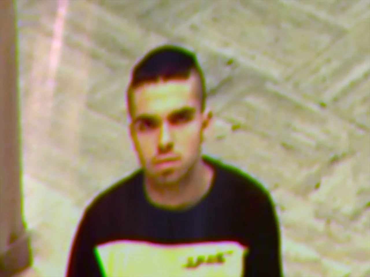 Violet (2014) movie still: medium — A stone-paved courtyard is captured from a high-angle security camera perspective. A young man stands in the…
