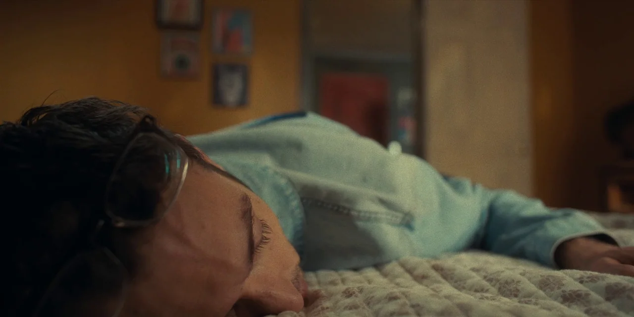 Destroy All Neighbours (2024) movie still: closeup — A dark bedroom features a person sleeping on a textured patterned blanket. Small framed wall…