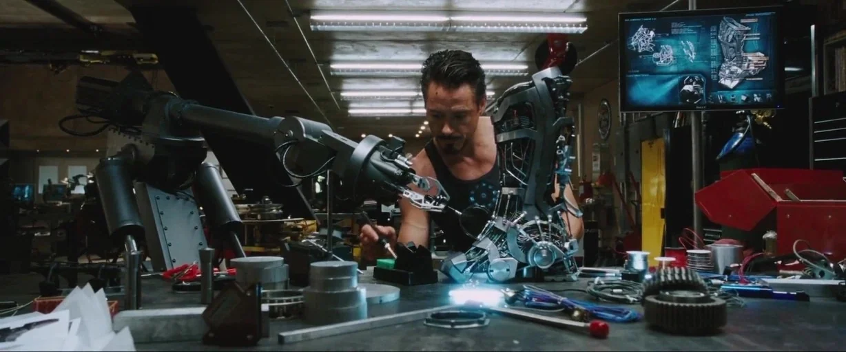 Iron Man (2008) movie still: medium — A cluttered high-tech workshop filled with mechanical parts and industrial robot arms. A man with a goatee…