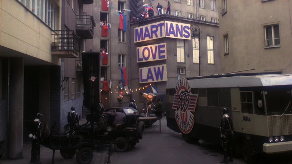 The War of the Worlds: Next Century (1981) movie still: wide — A dense urban courtyard features large posters reading 'MARTIANS LOVE LAW' on…