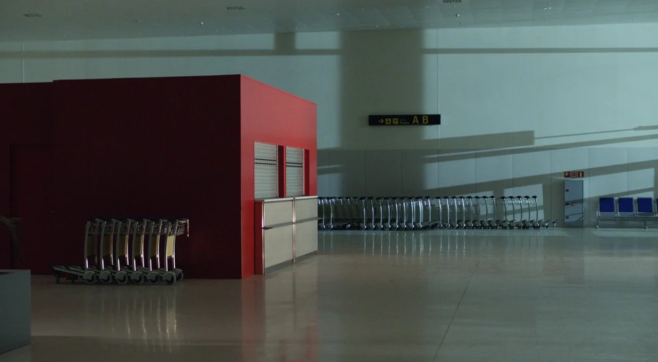 I’m So Excited! (2013) movie still: wide — A quiet airport terminal features a large red kiosk and rows of empty carts. Clean tile floors reflect…