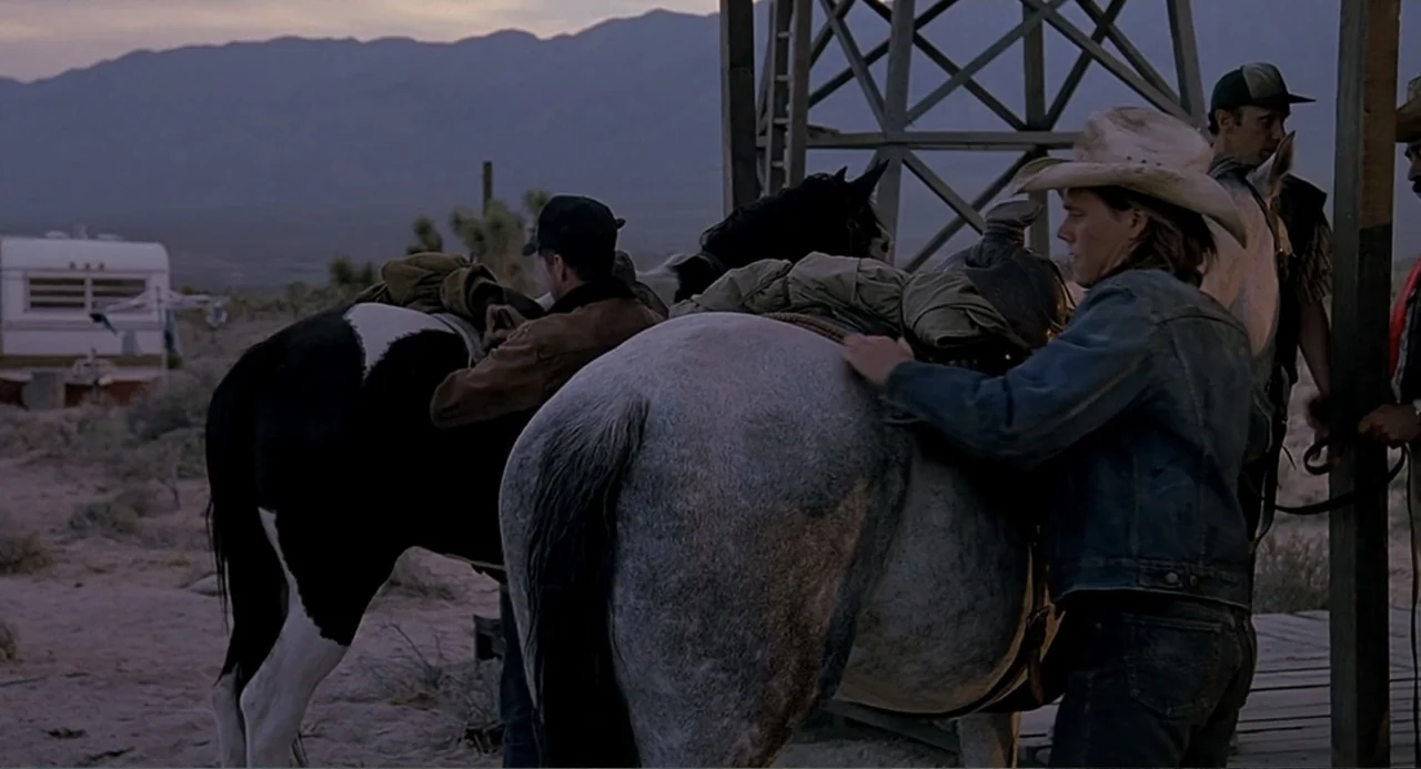 Tremors (1990) movie still: medium — A high desert ranch area with a wooden tower and distant mountains. Three men prepare two horses with saddles…
