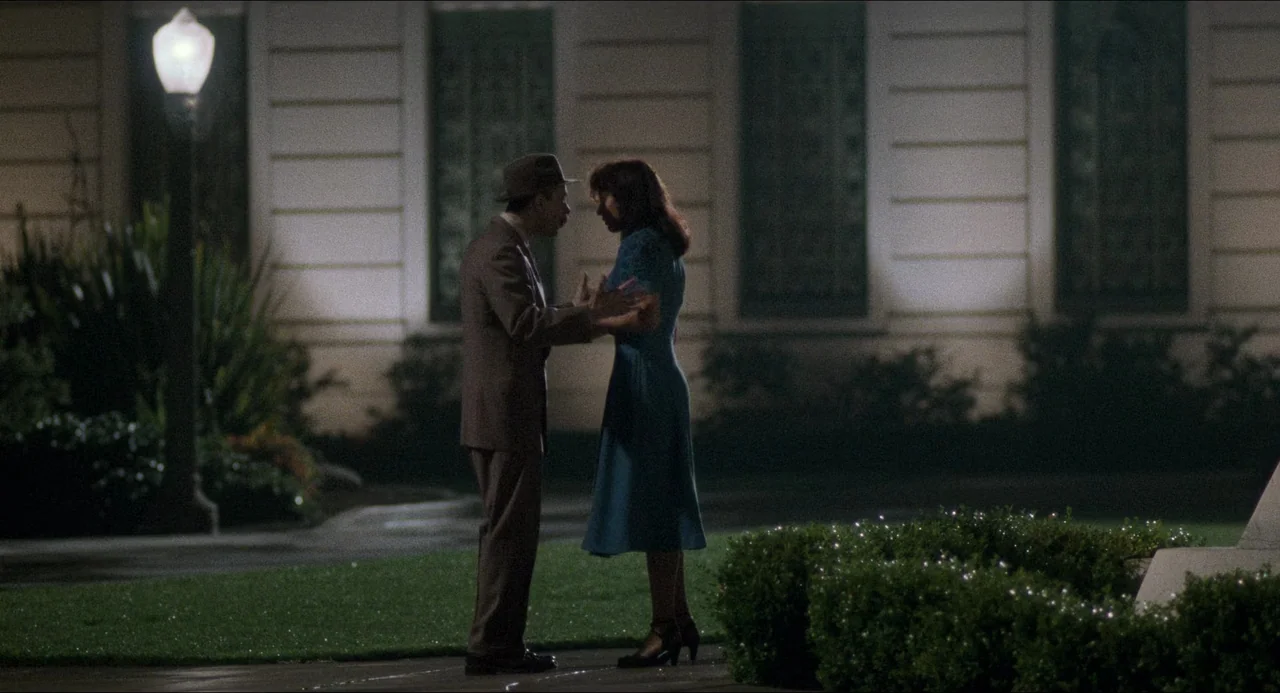 Devil In A Blue Dress (1995) movie still: fullbody — A dark outdoor area featuring a manicured lawn and a tall, glowing streetlamp. A man in a suit…