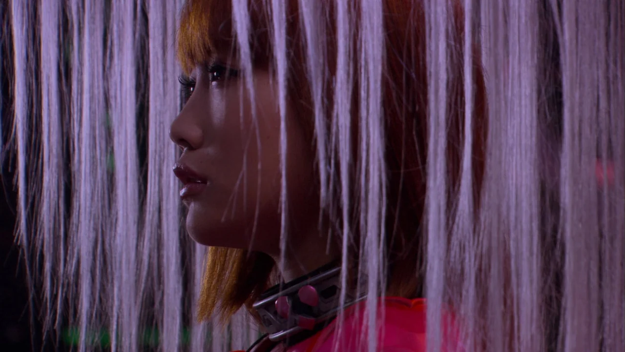 Cutie Honey (2004) movie still: closeup — A person stands behind hanging white fibers in a dark space. Purple light highlights her profile and…