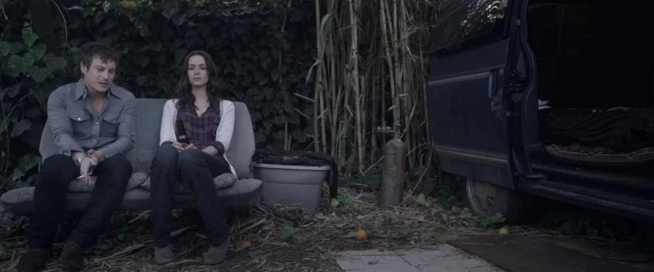 Starry Eyes (2014) movie still: wide — An outdoor area with a car bench seat placed amidst tall grass and vines. A man and a woman sit side by side…