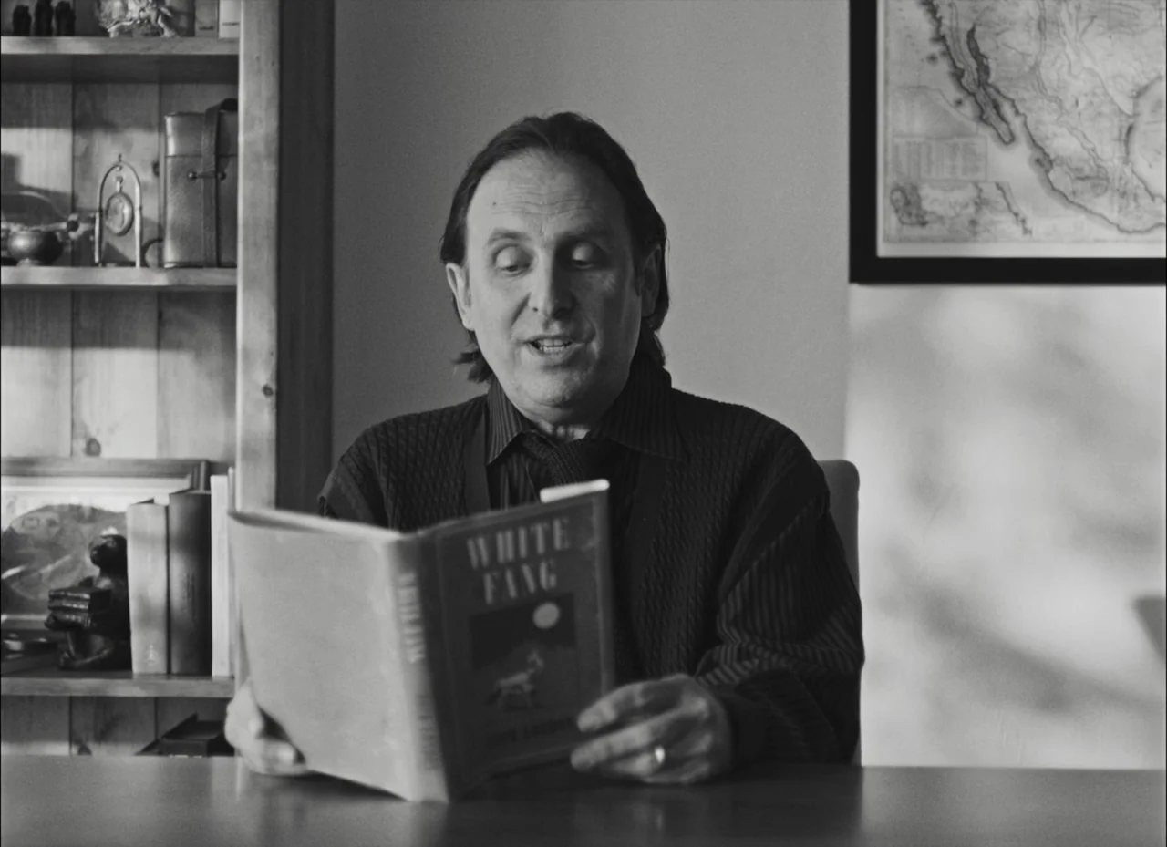 Fremont (2023) movie still: medium — A man sits at a desk in a room featuring a wooden bookshelf and map. He is reading the book White Fang while…